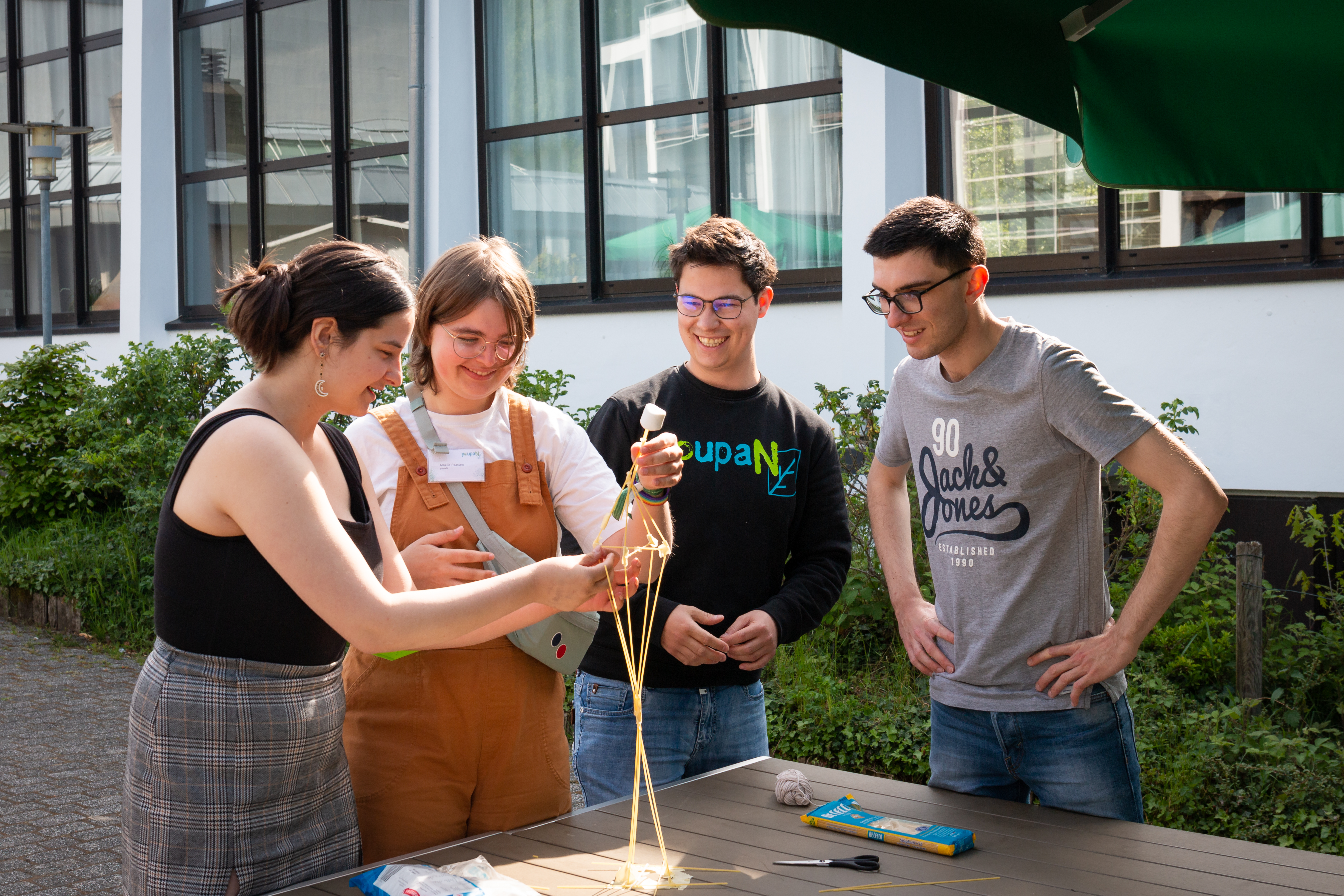 Vier Mitglieder des Jugendforums youpaN stehen im Freien um einen Tisch herum und haben aus Spaghetti und Marshmallows eine Art Skulptur gebaut.