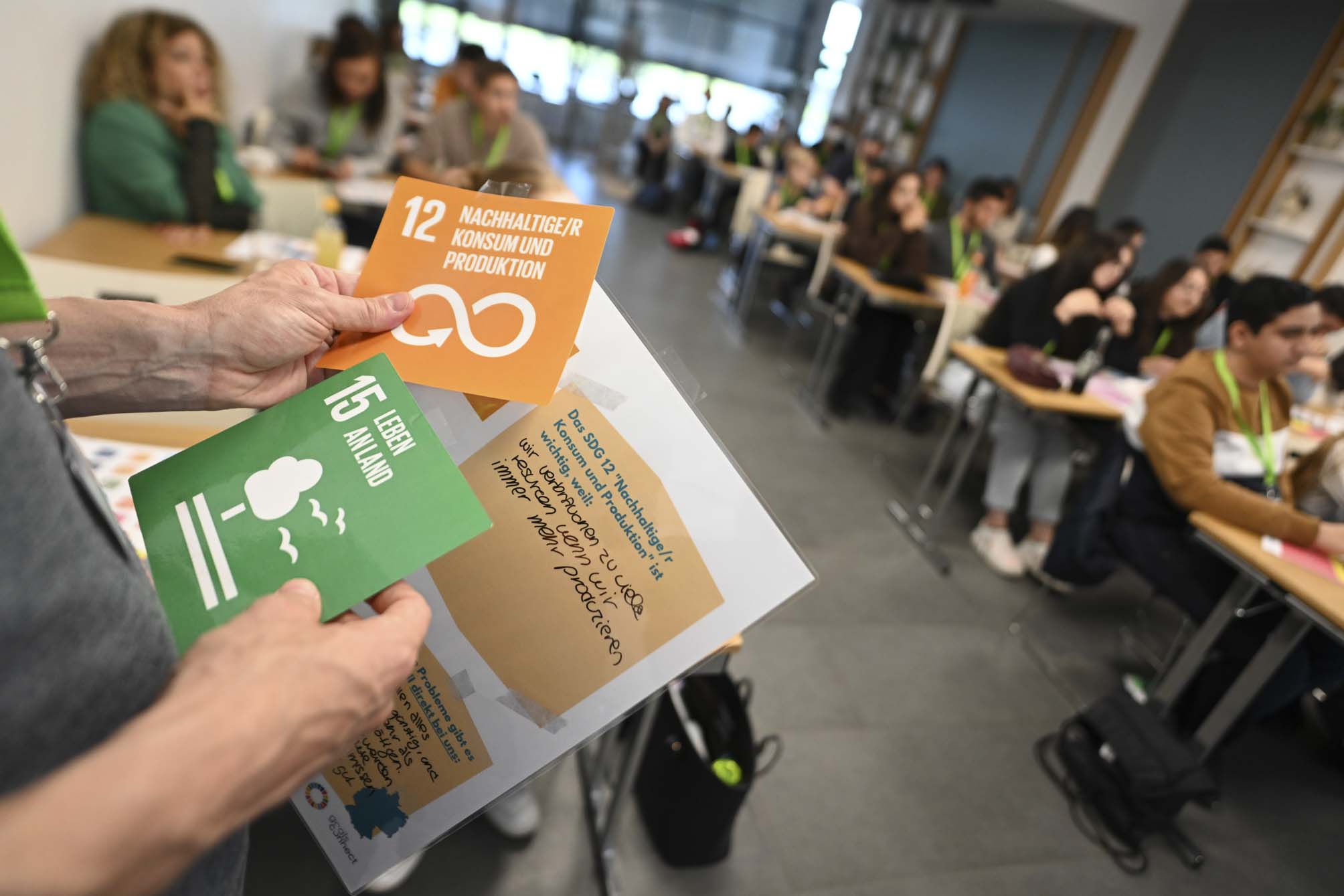 Eine Person hält zwei Karten in der Hand, die Nachhaltigkeitsziel 12 und 15 zeigen; im Hintergrund sitzen Jugendliche an Tischen