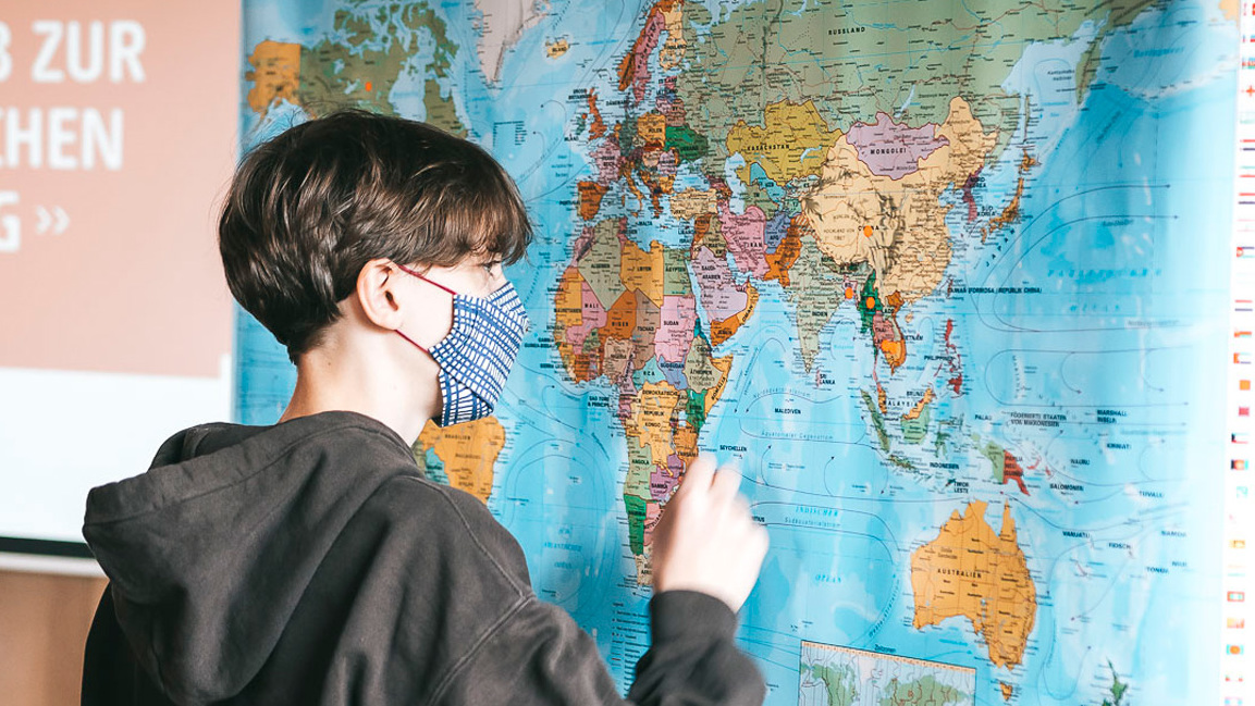 Ein Junge mit Maske steht vor einer Weltkarte.