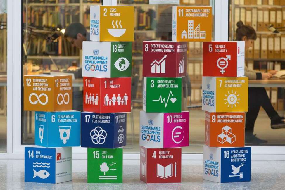Aufgestapelte, bunte Würfel, auf die die SDGs gedruckt sind vor zwei Personen, die im Hintergrund an Tischen sitzend auf aufgeschlagene Bücher schauen